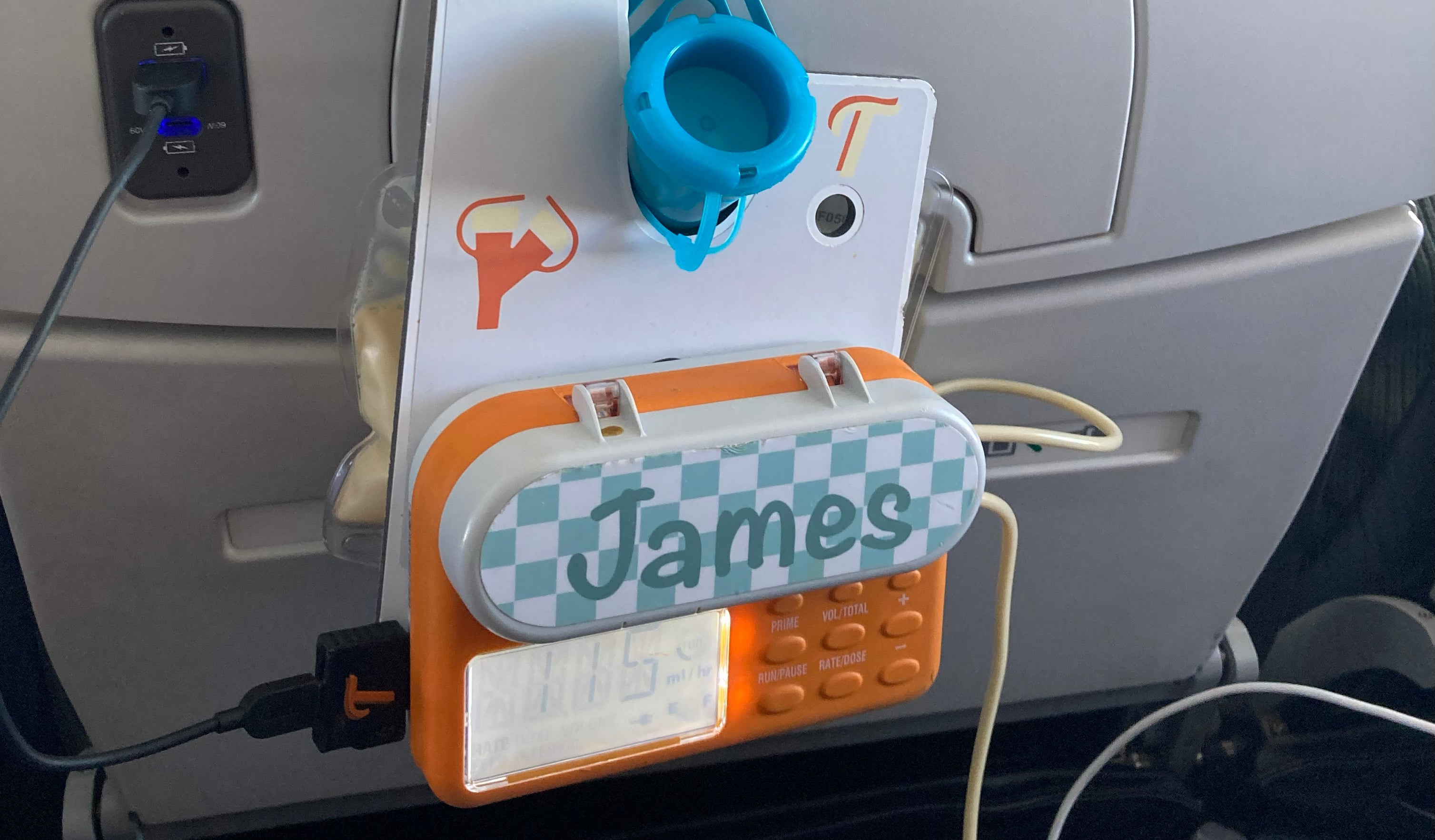 An orange Infinity pump attached to a pump hanger that is hanging from the back of an airplane seat. The pump is charging with a USB-C charging adapter that is plugged into the USB port of the plane seat