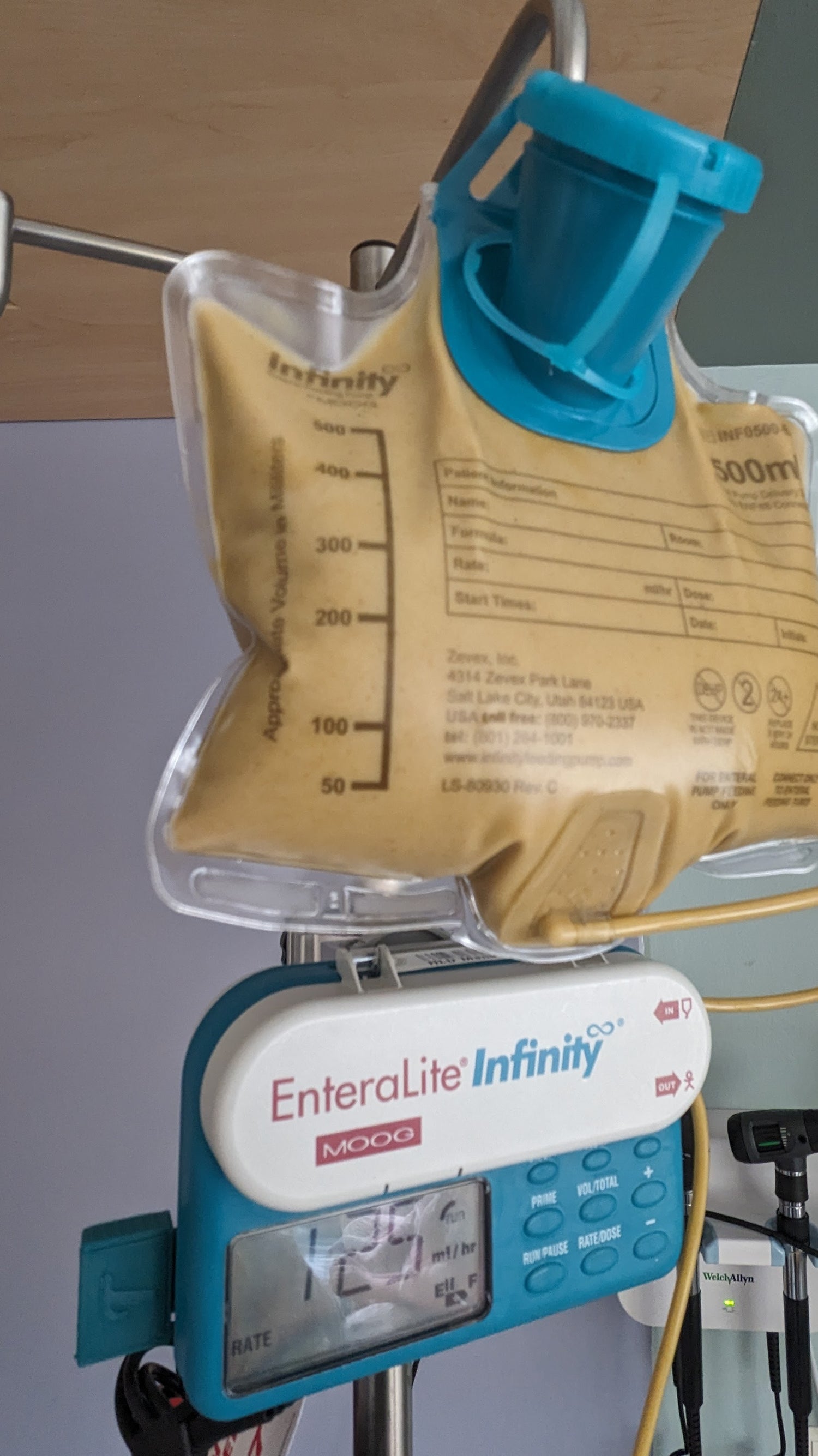 An enteral feeding pump with a tubie tech usb-c charger and full feeding bag hanging on an IV pole in a hospital room